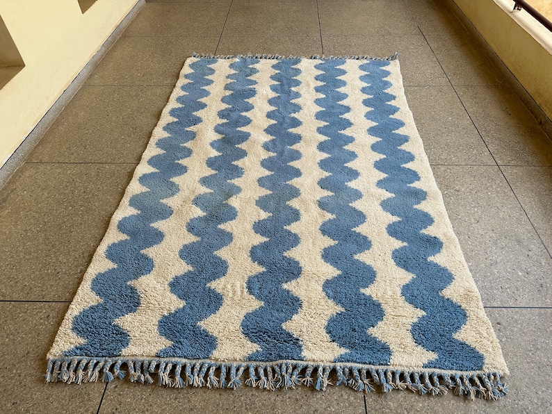 Pu&ograve; includere: Un tappeto rettangolare con sfondo color crema e strisce ondulate azzurre. Il tappeto ha una frangia su entrambe le estremit&agrave; corte. Il tappeto &egrave; realizzato in un materiale morbido e strutturato.