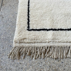 May include: A white rug with a black border and fringe. The rug is made of a soft, fluffy material.