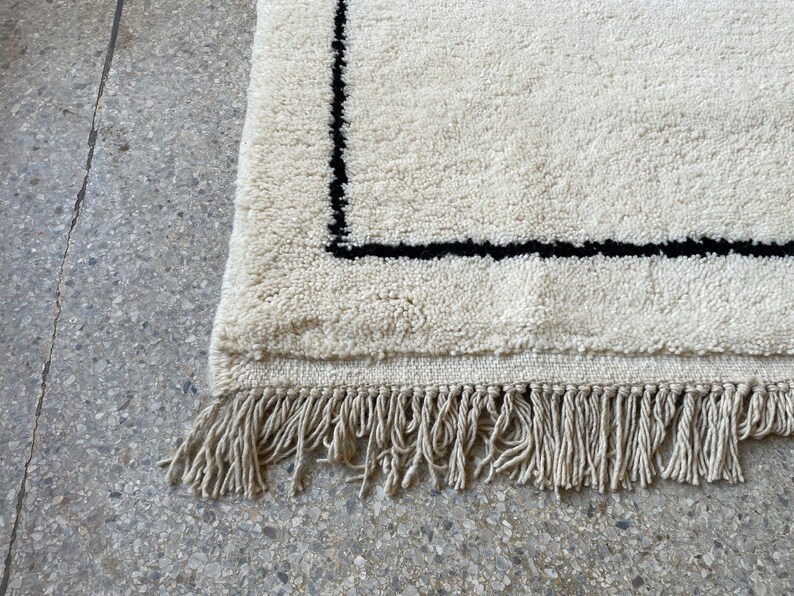May include: A white rug with a black border and fringe. The rug is made of a soft, fluffy material.