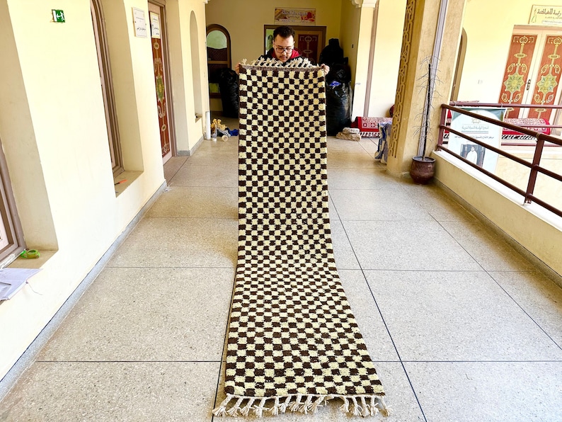 May include: A long, rectangular rug with a checkered pattern in brown and cream colors. The rug has fringe on the ends.