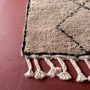 May include: A beige and black Moroccan rug with a diamond pattern and fringe. The rug is made of wool and has a soft, textured feel.