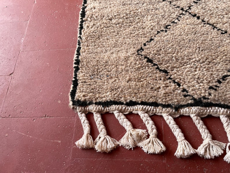 May include: A beige and black Moroccan rug with a diamond pattern and fringe. The rug is made of wool and has a soft, textured feel.