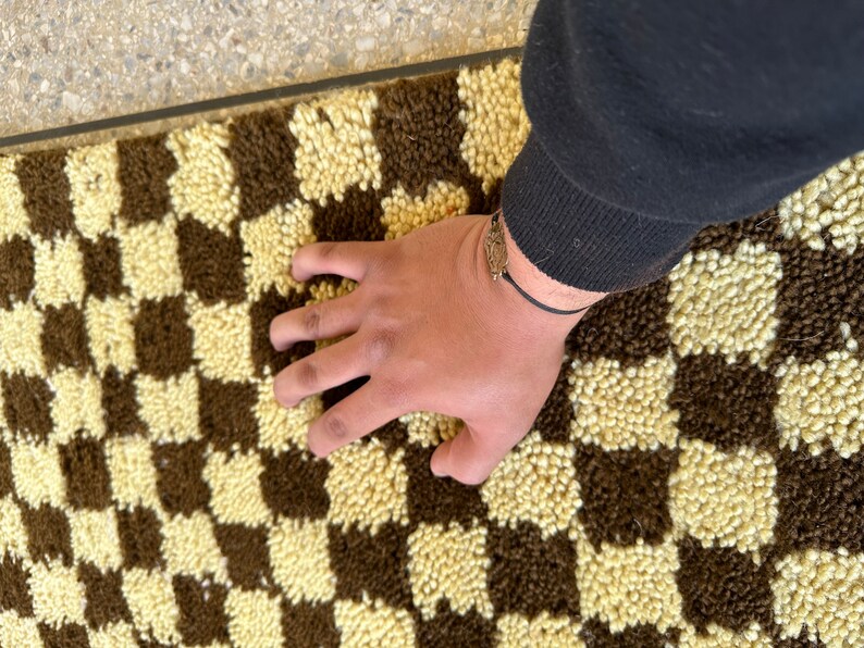 May include: A brown and cream checkered rug with a hand resting on the surface.