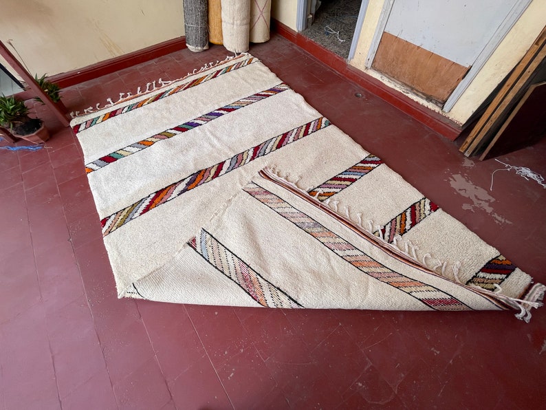 Puede incluir: Una alfombra blanca con rayas de colores tejidas en la tela. La alfombra est&aacute; doblada por la mitad y colocada sobre un suelo de madera.