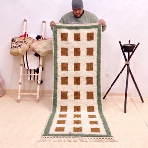 Peut inclure: Un tapis rectangulaire avec un fond crème et un motif de carrés marron. La bordure est verte. Le tapis est tenu par une personne. D'autres articles en arrière-plan comprennent une échelle avec des sacs et un support de trépied noir.