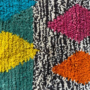 K&ouml;nnte beinhalten: Nahaufnahme eines bunten Teppichs mit geometrischem Muster. Der Teppich besteht aus verschiedenen farbigen Garnen, darunter Blau, Gr&uuml;n, Gelb, Rosa und Orange. Der Teppich hat eine strukturierte Oberfl&auml;che.
