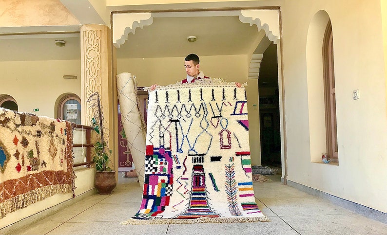 May include: A man holds up a white rug with colorful geometric patterns and designs. The rug is woven with wool and has a fringe border.