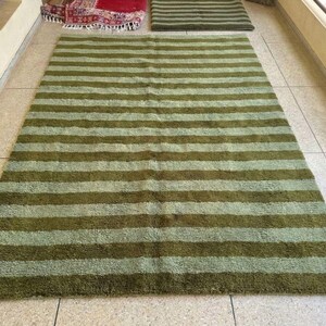 May include: A large, rectangular rug with horizontal stripes in shades of green and olive. The rug is made of a soft, textured material and is placed on a tiled floor. Additional rugs are visible in the background.