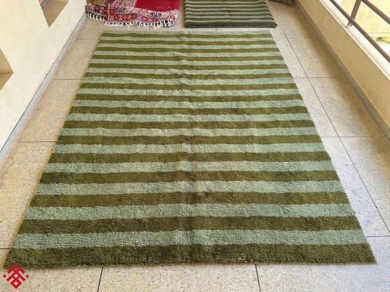May include: A large, rectangular rug with horizontal stripes in shades of green and olive. The rug is made of a soft, textured material and is placed on a tiled floor. Additional rugs are visible in the background.