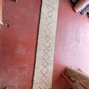 May include: A long, off-white runner rug with a black diamond pattern. The rug has fringe on the ends.