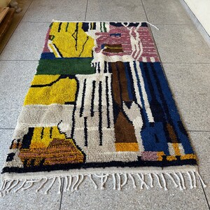 Puede incluir: Una alfombra abstracta colorida con un dise&ntilde;o geom&eacute;trico en tonos de amarillo, verde, azul, marr&oacute;n y rosa. La alfombra tiene un borde de flecos.