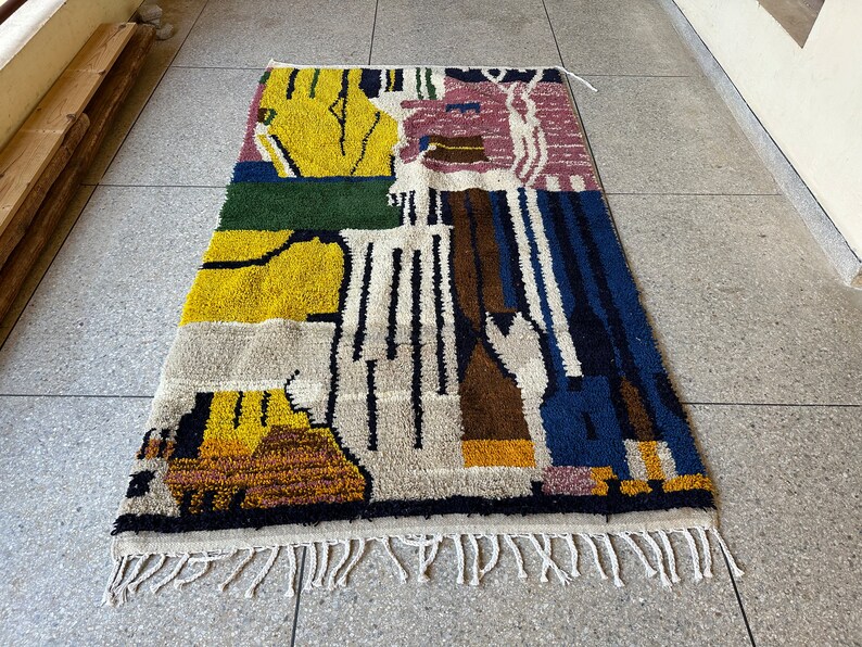 Puede incluir: Una alfombra abstracta colorida con un dise&ntilde;o geom&eacute;trico en tonos de amarillo, verde, azul, marr&oacute;n y rosa. La alfombra tiene un borde de flecos.