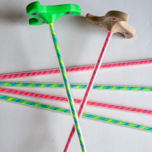 Pode incluir: Duas hastes longas e finas de plástico transparente com listras diagonais em cores neon rosa, azul e verde. Uma haste possui um cabo verde brilhante de três pontas, enquanto a outra tem um cabo bege com parafusos metálicos visíveis.