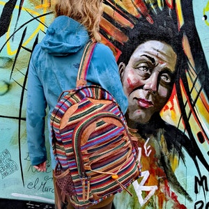 Op de afbeelding: Een vrouw met een blauwe capuchontrui en een kleurrijke gestreepte rugzak met leren accenten staat voor een graffitimuur. De muur heeft een grote, kleurrijke muurschildering met een gezicht.