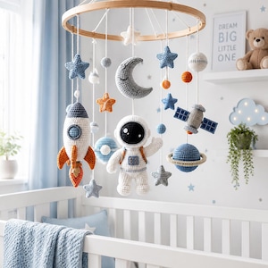 Può includere: Una giostrina per culla a tema spaziale con figure all'uncinetto tra cui un astronauta, un razzo, una luna crescente, un pianeta con anelli, un satellite e varie stelle nei toni del blu, bianco e arancione, sospese a un cerchio in legno.