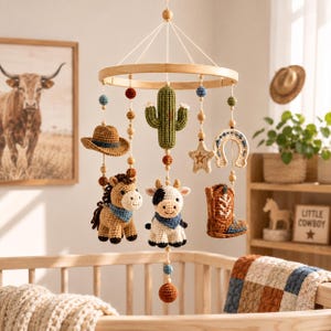 Op de afbeelding: Een gehaakte babymobiel met een houten ringframe en hangende garenfiguren, waaronder een bruin paard, een zwart-witte koe, een groene cactus, een bruine cowboyhoed, een beige ster, een hoefijzer en een bruine cowboylaars met blauwe accenten.