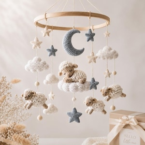 May include: A hanging nursery mobile featuring crocheted figures of three sheep, a crescent moon, several stars, and clouds suspended from a circular wooden frame. The sheep are cream-coloured, the moon and some stars are blue, and others are cream.