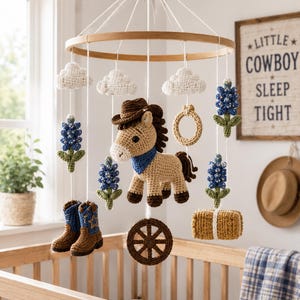May include: A crochet baby mobile featuring a central tan horse wearing a brown cowboy hat and blue bandana, surrounded by hanging elements including white clouds, blue flowers, brown cowboy boots, a tan lasso, a brown wagon wheel, and a hay bale.