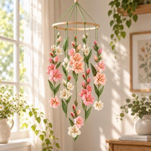 Puede incluir: Un móvil colgante que presenta flores de gladiolo tejidas a ganchillo en tonos rosa, melocotón y blanco, unidas a tallos y hojas verdes de ganchillo. Las hebras florales cuelgan de un aro de madera circular mediante cordones de hilo verde.