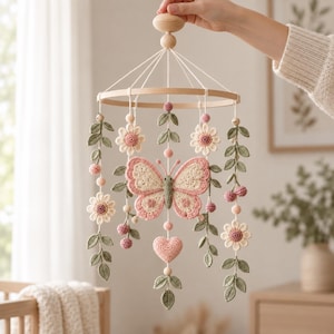 May include: A hanging mobile featuring a central crocheted butterfly and heart, surrounded by crocheted flowers, leaves, and small spheres in shades of pink, cream, and sage green, all suspended from a circular wooden frame with white strings.