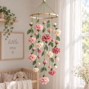 Puede incluir: Un móvil colgante que presenta un marco circular de madera con múltiples hilos colgantes de flores y hojas de ganchillo en tonos rosa, crema y verde. Los elementos florales incluyen flores completamente abiertas y pequeños capullos cerrados.