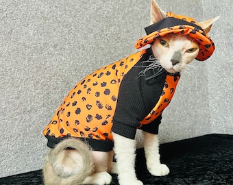 May include: A Sphynx cat wearing an orange and black patterned outfit and matching hat. The outfit has a black ribbed collar and sleeves, with an orange body covered in black spots. The hat has a black band. The cat is sitting on a black surface.