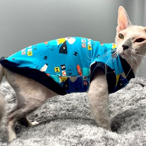 Peut inclure: Un chat sans poil portant une chemise bleue et noire avec un imprimé graphique coloré. La chemise porte le texte "Rock Star". Le chat est debout sur une surface moelleuse grise.