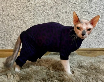 Puede incluir: Un gato sin pelo con un mono de leopardo morado y negro. El gato está de pie sobre una alfombra peluda de color marrón claro.