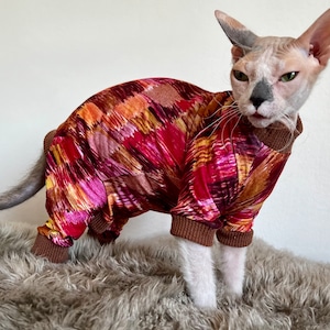 May include: A Sphynx cat wearing a colorful jumpsuit. The jumpsuit features a vibrant abstract pattern in shades of red, pink, orange, and brown. It has brown ribbed cuffs and a matching collar. The cat is standing on a gray, fluffy surface.