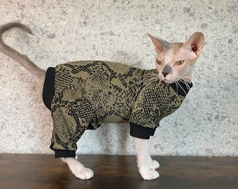 May include: A hairless cat wearing a green and black snake print jumpsuit. The cat is standing on a wooden surface.