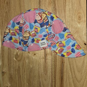 May include: A light blue welding cap featuring a repeating pattern of Dubble Bubble gum wrappers, pink bubble shapes, and cartoon characters. A white rectangular fabric label with the text Hollywood Welding Hats is stitched onto the side.