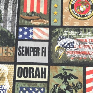 May include: A fabric panel featuring a collage of military-themed imagery. The design includes American flags, the United States Marine Corps emblem, stars, and phrases like "Semper Fi" and "Oorah." The color palette is dominated by red, white, blue, and olive green.