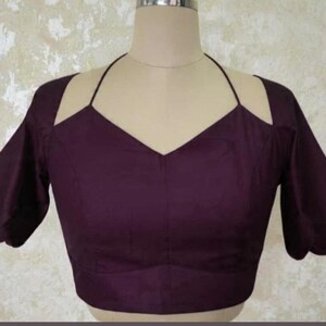 May include: A plum-coloured blouse with a halter neckline and cut-out shoulder design. The short sleeves have a scalloped edge. The blouse is displayed on a mannequin.
