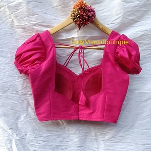 May include: A bright pink, sleeveless blouse with puffed sleeves and a low back. The blouse has a tie at the back and the text "DolyManviBoutique" is printed on the hanger.