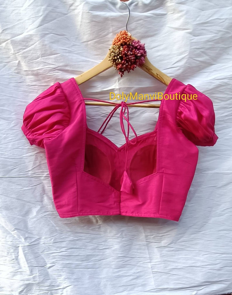 May include: A bright pink, sleeveless blouse with puffed sleeves and a low back. The blouse has a tie at the back and the text "DolyManviBoutique" is printed on the hanger.