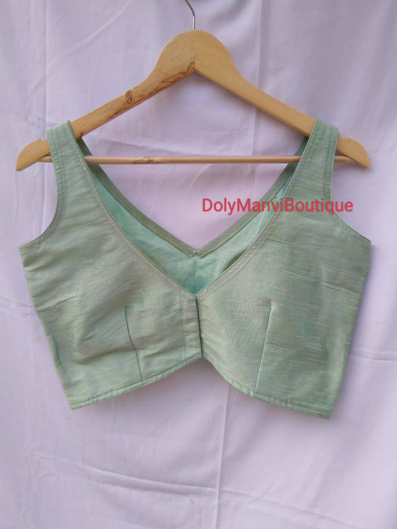 May include: A light green sleeveless blouse with a V-neckline. The blouse is made of a soft, silky fabric and has a simple, elegant design. The text "DolyManviBoutique" is visible on the blouse.