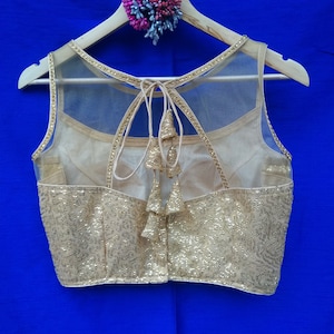 May include: A gold sequined sleeveless blouse with a sheer mesh back and a tie closure. The blouse has a scalloped neckline and is decorated with small gold beads.