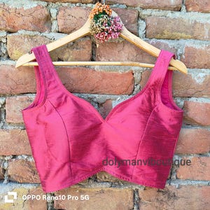 May include: A magenta-colored silk blouse with a V-neck design, displayed on a wooden hanger. The blouse has a smooth texture and is hanging against a brick wall. The text "dolymanviboutique" is visible on the blouse.