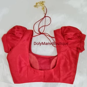 May include: Red sleeveless blouse with puff sleeves and a low back. The blouse has a tie closure at the back of the neck. The text "DolyManviBoutique" is printed on the front of the blouse.