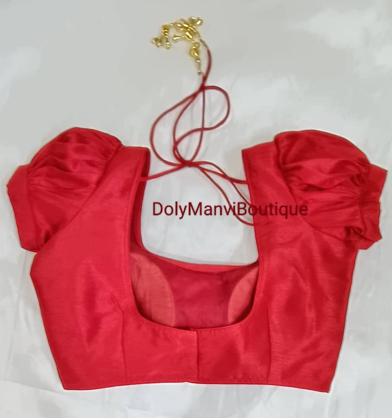 May include: Red sleeveless blouse with puff sleeves and a low back. The blouse has a tie closure at the back of the neck. The text "DolyManviBoutique" is printed on the front of the blouse.