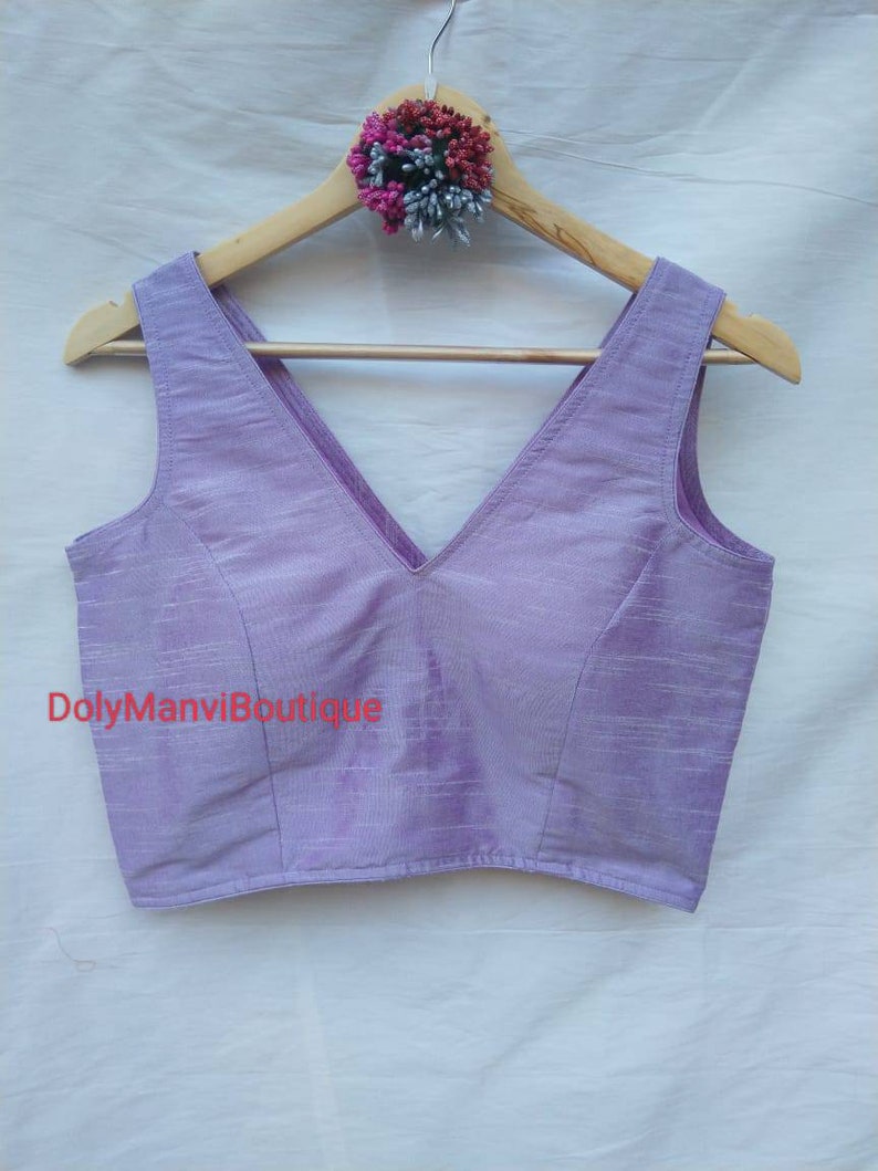 May include: A lavender sleeveless blouse with a deep V-neckline. The blouse is made of a soft, silky fabric and has a subtle sheen. The text "DolyManviBoutique" is printed on the front of the blouse.