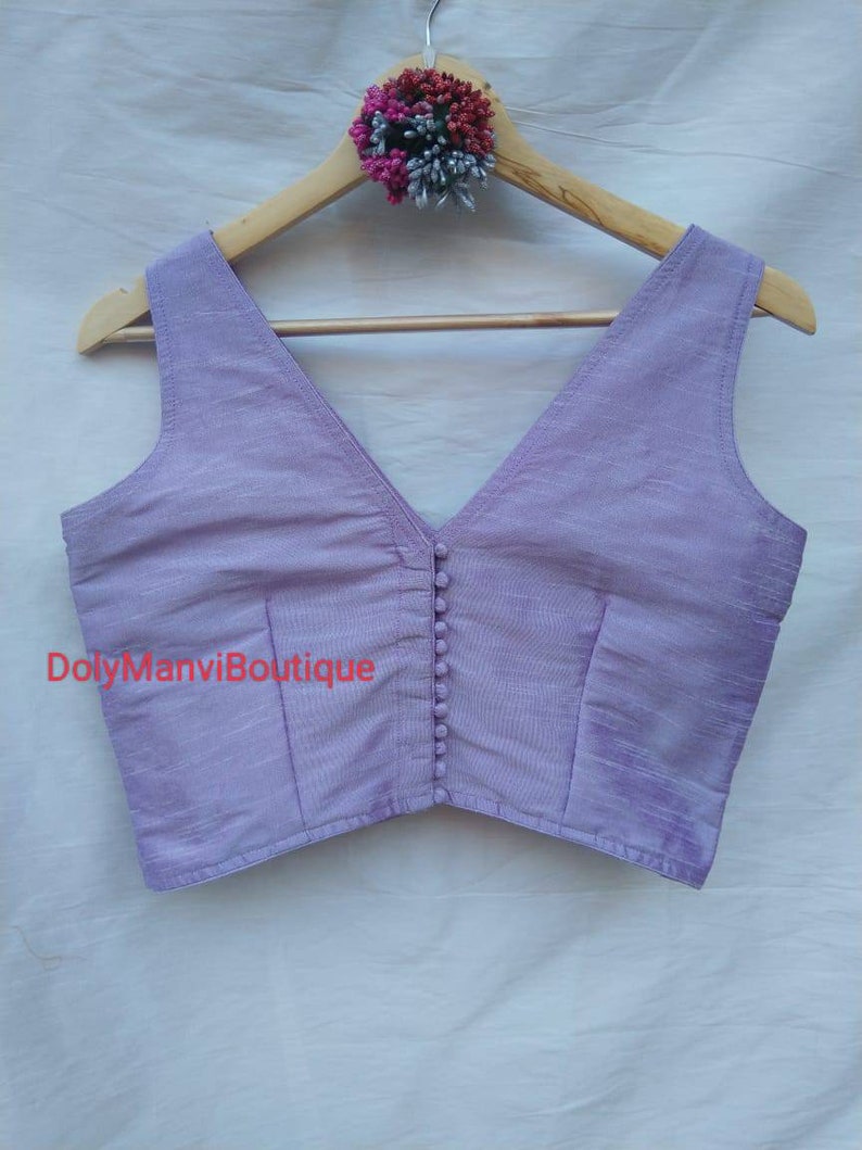 May include: A lavender sleeveless blouse with a deep V-neckline and button closure. The blouse is made of a soft, silky fabric and has a cropped fit.