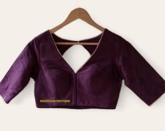 May include: A deep purple blouse with elbow-length sleeves, a V-neckline, and a triangular cutout at the back. The neckline is trimmed with gold piping. The text DOLYMANVIBUTIQUE is printed in yellow on the lower left side of the garment.