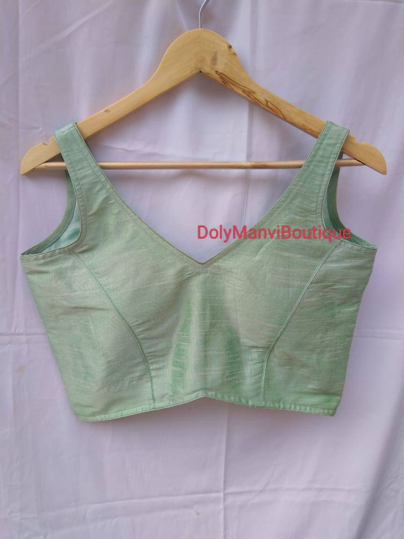 May include: A light green sleeveless blouse with a V-neckline, hanging on a wooden hanger. The blouse has a subtle sheen and is made of a soft, flowing fabric.