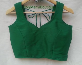 Puede incluir: Una blusa sin mangas de color verde esmeralda con cuello en V y cierre de lazo en la espalda. La blusa está colgada en una percha de madera con un adorno floral. El texto "DolyManviBoutique" es visible en la percha.