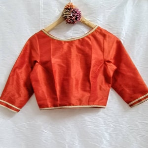 Puede incluir: Una blusa de color óxido con mangas tres cuartos, cuello redondo y ribetes dorados. La blusa está colgada en una percha de madera.