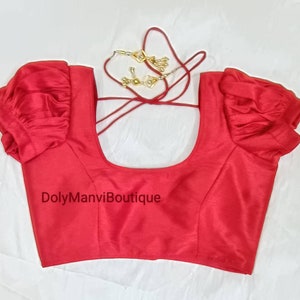 May include: Red sleeveless blouse with puffed sleeves and a scoop neck. The blouse has a tie closure at the back. The text "DolyManviBoutique" is printed on the front of the blouse.