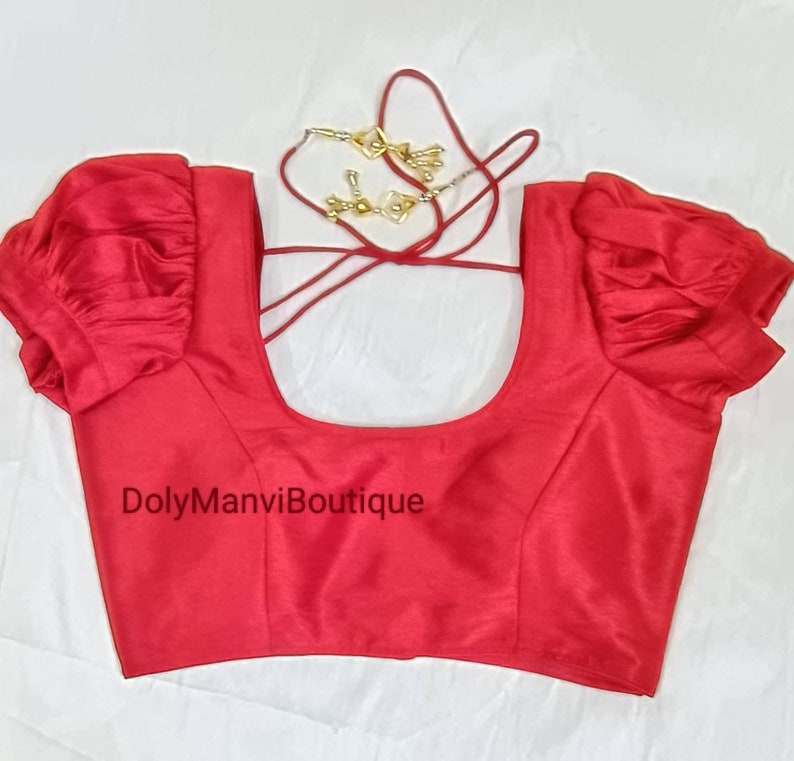 May include: Red sleeveless blouse with puffed sleeves and a scoop neck. The blouse has a tie closure at the back. The text "DolyManviBoutique" is printed on the front of the blouse.