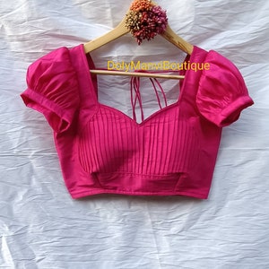 May include: A pink, pleated, sleeveless blouse with puffed sleeves and a sweetheart neckline. The blouse is hanging on a wooden hanger with a floral embellishment.