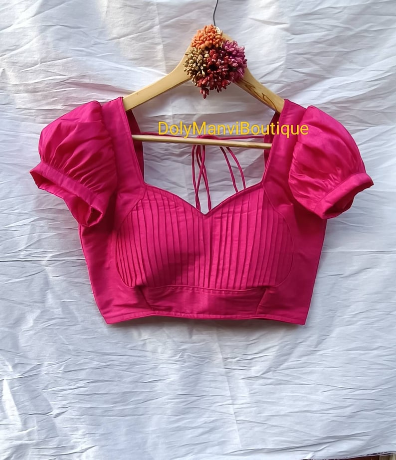 May include: A pink, pleated, sleeveless blouse with puffed sleeves and a sweetheart neckline. The blouse is hanging on a wooden hanger with a floral embellishment.